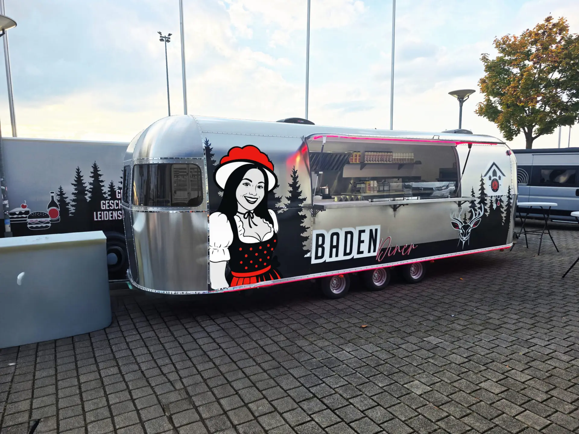 Silberner Retro-Verkaufswagen mit der Aufschrift „BADEN Diner“. Auf der Seite ist eine Grafik einer lächelnden Frau in Tracht zu sehen. Der Wagen steht auf einem gepflasterten Platz, die Verkaufsklappe ist geöffnet und gibt den Blick auf die Küche frei.