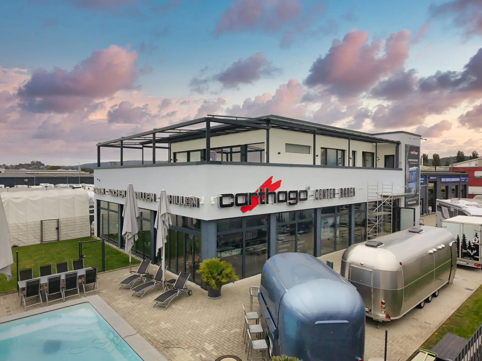 Eine Drohnenaufnahme des Carthago Centers Baden. Ein weißes, zweistöckiges Gebäude mit der Aufschrift „Carthago Center Baden“. Im Vordergrund ein Außenbereich mit Pool, anthrazitfarbenen Liegestühlen und markanten, silbernen Retro-Wohnwagen auf einem gepflasterten Vorplatz.